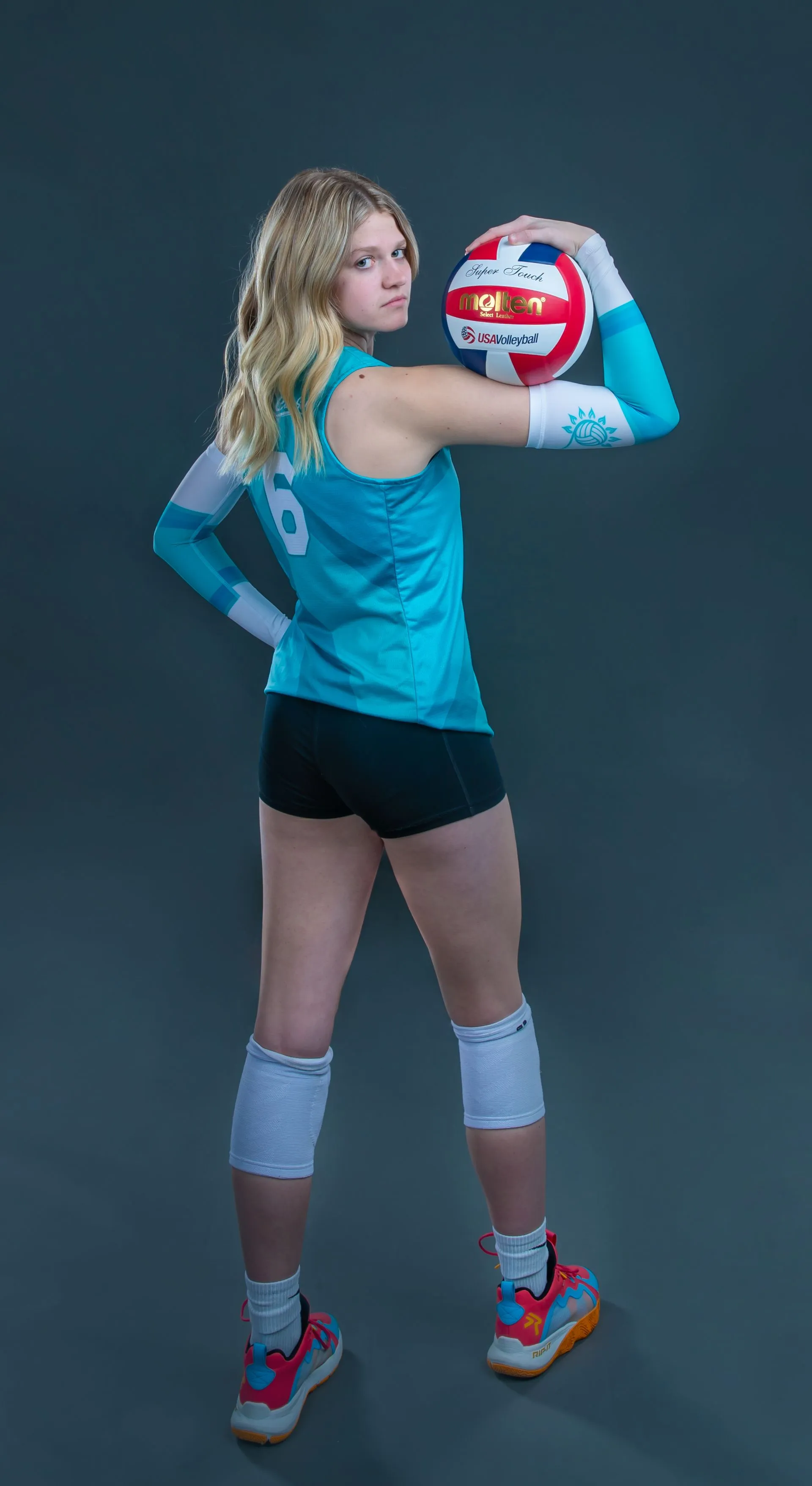 Athlete in turquoise volleyball gear holds a red-white ball against a dark background.