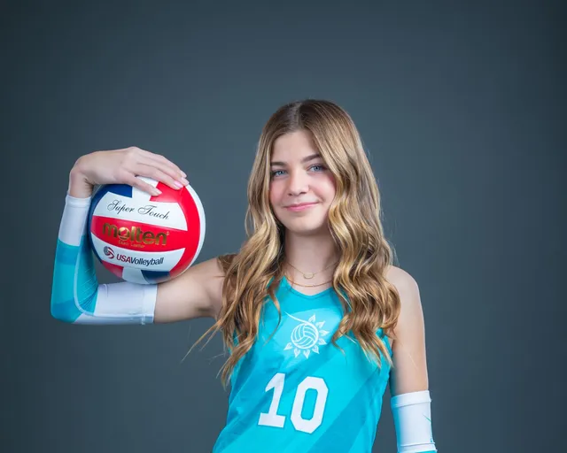 Girl in teal volleyball jersey holding a red-and-white volleyball and smiling against a dark background